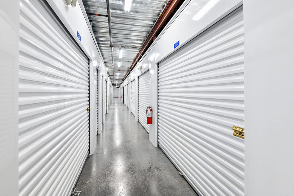Long indoor corridor with white metal roll-up storage unit doors on both sides, polished concrete floor, fluorescent ceiling lights, and red fire extinguisher mounted on wall.