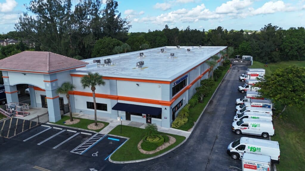 Aerial view of U-Haul self-storage facility with white building, orange trim, flat roof with HVAC units, and fleet of rental trucks parked along driveway.