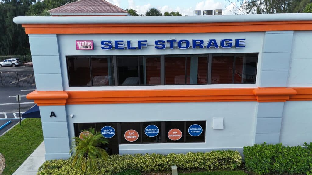 Value Store It self-storage facility exterior with light blue walls, orange trim, and entrance signage advertising truck rentals, A/C units, referral program, no deposit, and internet access.