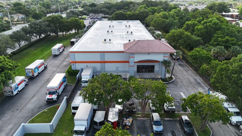 Aerial view of U-Haul self-storage facility with white building, red-trimmed roof, multiple rental trucks parked outside, and surrounding trees.