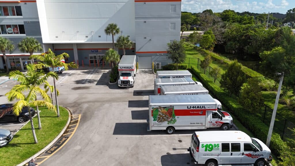 Aerial view of U-Haul rental trucks and moving vans parked in front of a modern self-storage facility with palm trees and green landscaping.