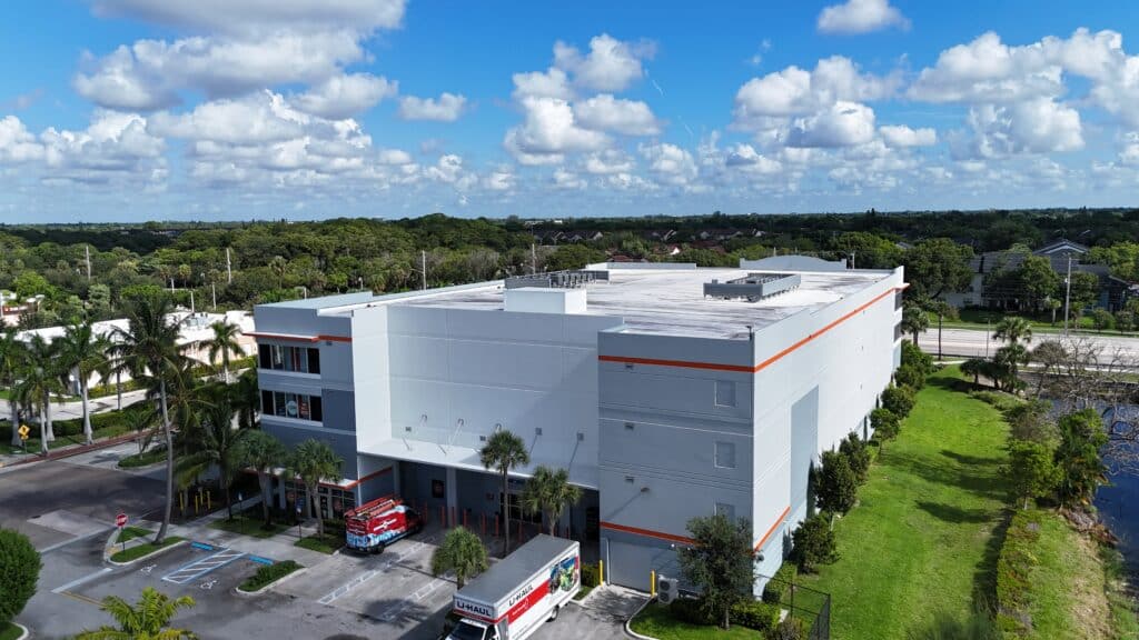 Aerial view of modern white and orange self-storage facility with flat roof and ground-level drive-up access, surrounded by palm trees and green landscape in Florida.