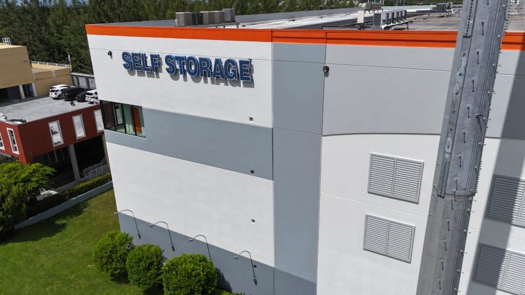 Exterior of a modern self-storage facility with white and gray paneling, orange trim, and blue "SELF STORAGE" signage, adjacent red office building visible on left.